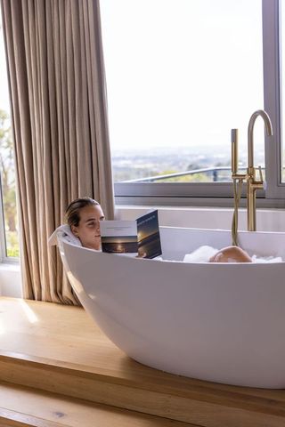 Woman relaxing in freestanding tub reading book by window overlooking city view