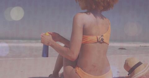 Woman in Orange Bikini Sitting on Beach Holding Sunscreen Facing Ocean