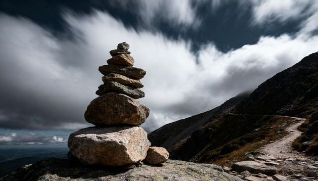 Balancing cairn on alpine mountain ridge with dramatic clouds and rugged footpath