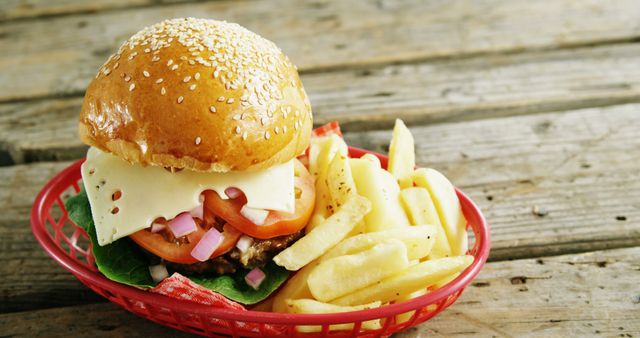 A cheeseburger with lettuce, tomato, onions, and a side of French fries is presented in a red basket on a wooden table. The meal represents a classic American fast-food option, often enjoyed for its satisfying taste and quick service.