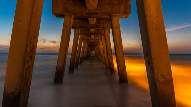 Long exposure photo showing the underside of a pier at sunset. The smooth, blurred effect of the waves creates a serene and tranquil atmosphere, making the pillars and sunset hues vibrant. Useful for backgrounds in presentations, nature themes, and travel industry promotions.