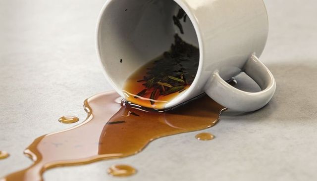 White ceramic mug tipping and spilling amber tea with loose leaves across light textured tabletop