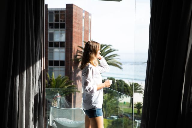 Side view close up of a young Caucasian woman wearing a white shirt standing on a balcony holding a cup of coffee and looking out, sea and beach in the background