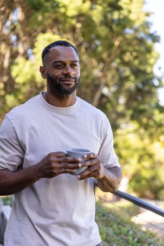 African american man standing on sunlit balcony holding mug, contemplative morning portrait
