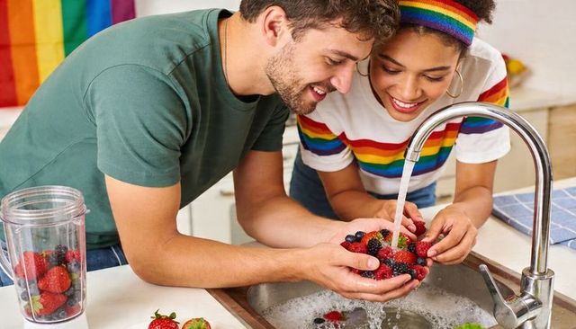 Couple washing mixed berries at sink with rainbow pride flag in kitchen