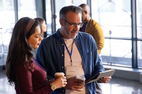 Colleagues reviewing tablet and holding coffee during conference networking with lanyards