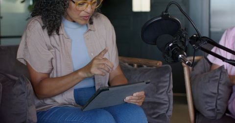 Woman hosting podcast with tablet pointing and professional microphone on living room sofa