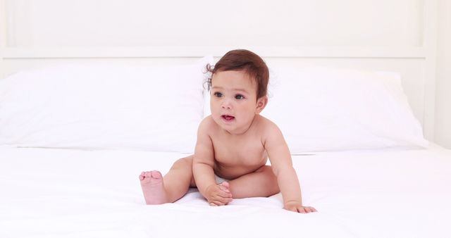 Baby sitting on white bed, looking curious and playful. Perfect for use in parenting blogs, childcare advertisements, baby product promotions, and educational materials.