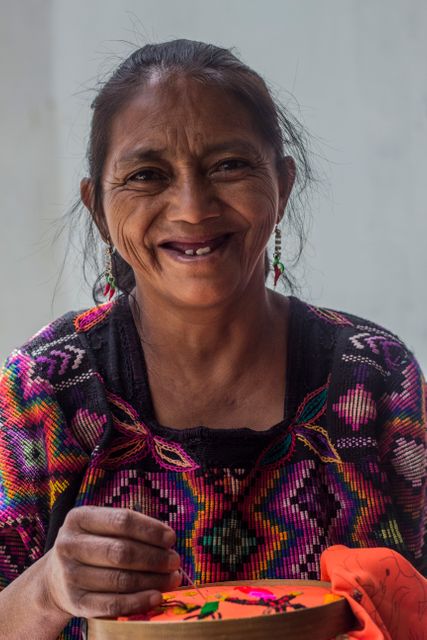 Indigenous woman smiling while embroidering traditional colorful textile. Ideal for topics related to cultural heritage, craftsmanship, traditional clothing, and artisan works. Suitable for educational content, promotion of folk art, artisanal fair advertisements, and cultural diversity.