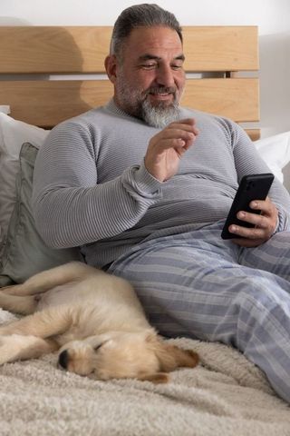 Senior man relaxing in bed using smartphone with sleeping golden retriever puppy nearby