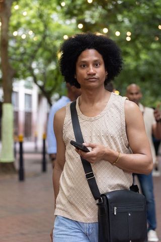 African american male walking tree-lined urban street holding smartphone and crossbody bag