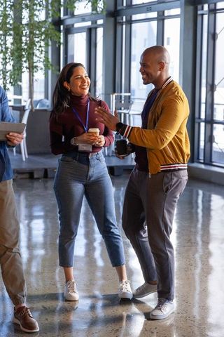 Diverse coworkers laughing and chatting with coffee and lanyards in modern office lounge