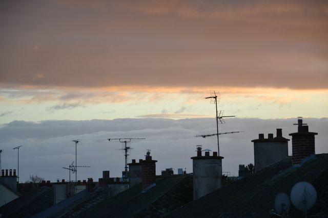 Sunset over rooftops showing TV antennas silhouetted against a cloudy sky. Useful for urban lifestyle, cityscape themes, or illustrating evening time concepts. Can be utilized in blogs, articles, or websites related to everyday urban living or discussions on technology and cityscapes.