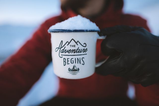 Person holding a white enamel mug filled with snow outdoors in a mountainous, winter landscape. Mug has a motivational quote on it, making it perfect for content related to adventure, inspiration, travel, and explorations. Ideal for use in blog posts, social media updates related to winter activities, travel adventures, and motivational content.