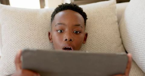 African American boy watching tablet closely while reclining on cream couch cushions