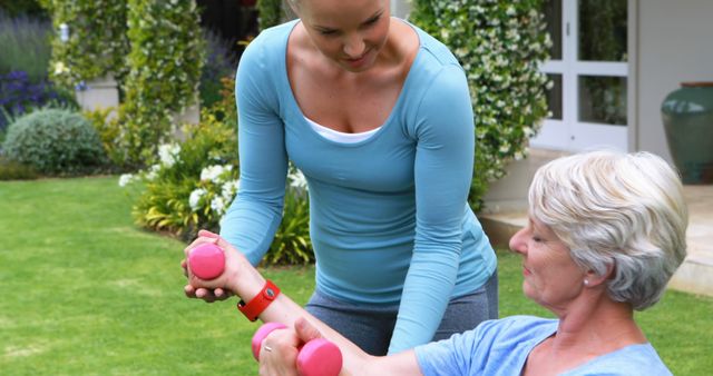 A personal trainer is assisting a senior woman with dumbbell exercises in a beautiful garden setting. This image highlights the importance of staying active as one ages and the benefits of personal coaching. Can be used for promoting fitness programs for seniors, physical therapy ads, healthy lifestyle content, or personal training services.