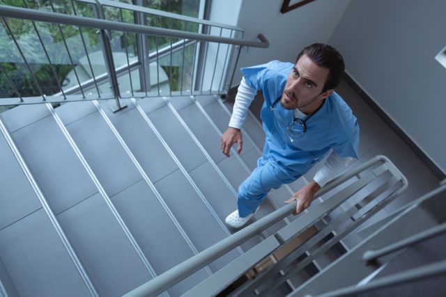 Male surgeon in blue scrubs walking up stairs in a hospital. Ideal for illustrating healthcare professionals, hospital environments, and medical facilities. Can be used in articles, brochures, and websites related to healthcare, medical staff, and hospital operations.