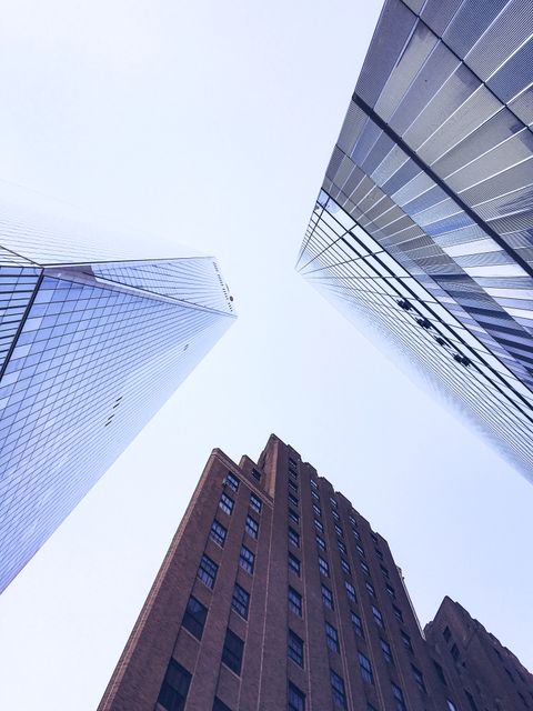 This image shows a low-angle view of modern high-rise buildings against a clear, blue sky. The buildings, with their glass facades and sleek designs, create an impressive cityscape. Ideal for use in urban development, real estate presentations, architecture portfolios, and city living advertisements.