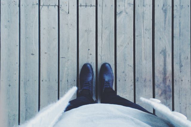 Person is standing on a wooden deck shown from an aerial perspective. Focus is on black shoes and worn planks of wood, creating contrast in texture. Great for themes of walking trails, outdoor fashion, lifestyle blogging, rustic environmental settings, or foot journey narratives.