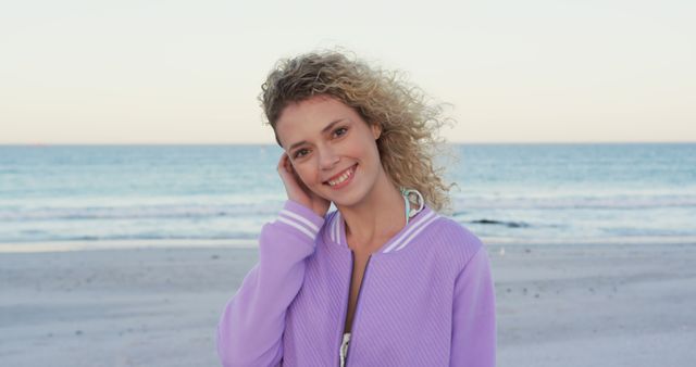Young woman with curly hair smiling while standing at a beach during sunset. She wears a casual purple jacket, and the ocean and sky provide a serene backdrop. Ideal for use in lifestyle blogs, travel websites, summer fashion promotions, and wellness articles.