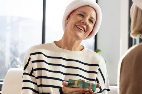 Senior woman wearing santa hat smiling while giving green gift on cozy sofa, joyful holiday moment