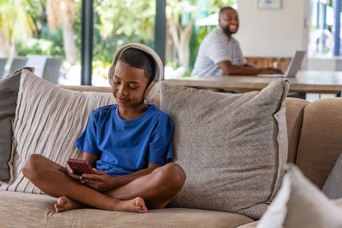 African american father working from home while son wearing headphones using smartphone