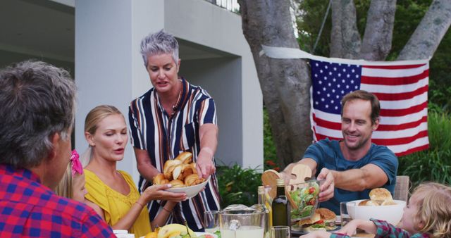 Family enjoying outdoor lunch on 4th of July, American flag in background. Perfect for depicting holiday gatherings, summer celebrations, American traditions.