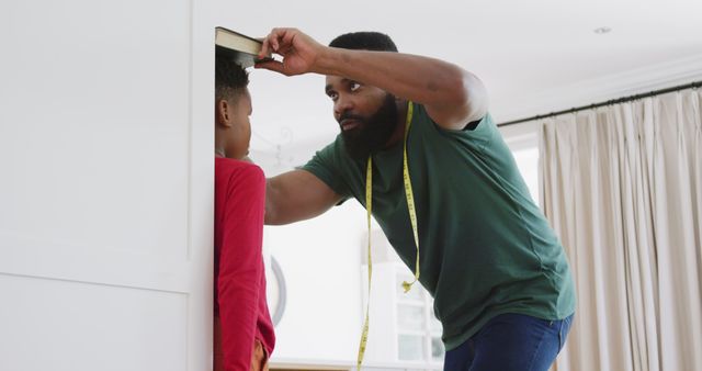 Father is checking growth by measuring son's height against wall using a book. Great for illustrating parent-child relationships, growth milestones, and family activities. Ideal for articles on parenting, child development, and routines at home.