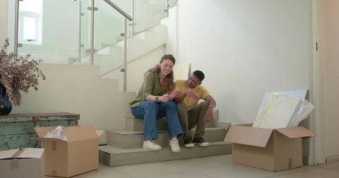 Laughing mixed-race couple unpacking moving boxes on foyer stairs, cozy new home moment