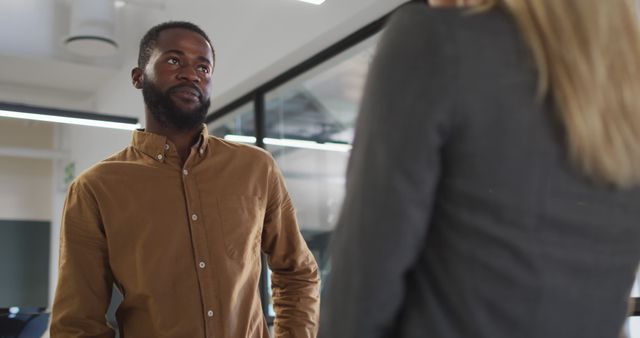 Man in brown shirt engaging in a casual discussion with a colleague in a contemporary office environment. The image captures the essence of professional communication and interaction among coworkers. Ideal for use in business-related articles, marketing materials, and presentations highlighting teamwork, collaboration, and modern work culture.