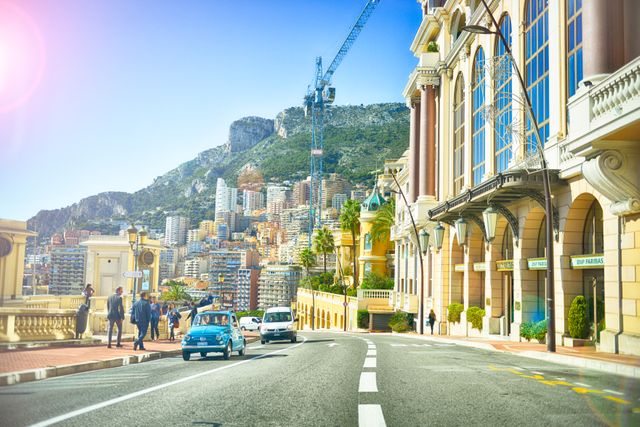 Features a vintage car driving on a street in Monte Carlo under a bright sunny sky. Modern buildings and luxurious architectures fill the background with a mountain view. Ideal for travel brochures, tourism advertisements, and lifestyle blogs focused on luxury travel and European cities.