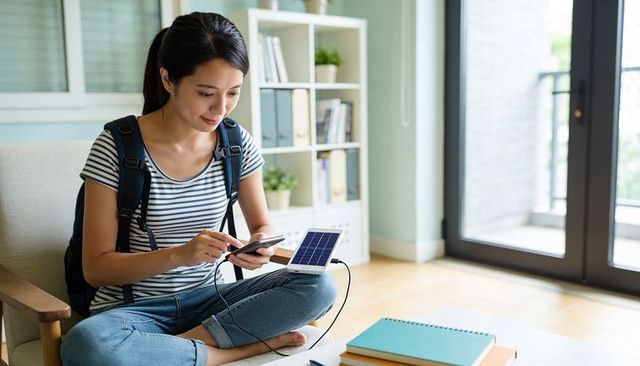 Woman charging smartphone with portable solar panel while studying at home, backpack and notebooks