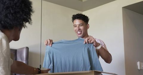 Smiling man holding light blue t-shirt while unpacking cardboard box in cozy home interior dining ar