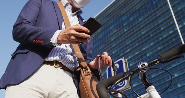 Asian man wearing face mask using smartphone while locking his bicycle at corporate park. health protection and safety during covid-19 pandemic concept