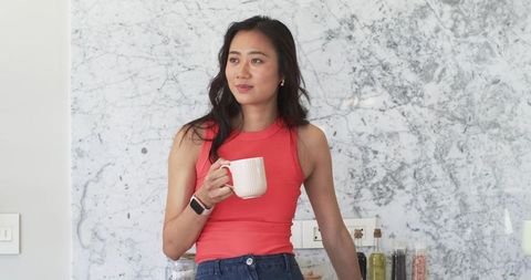 Asian woman holding coffee mug and leaning on marble kitchen counter with serene minimalist vibe