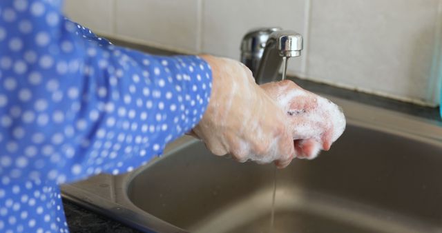 Person washing hands with soap under running water in kitchen or bathroom sink. Foaming soap visible. Can be used for promoting good hygiene, health safety tips, handwashing routines, or sanitary practices.