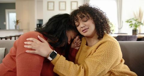Young women embracing on couch comforting friend in mustard sweater during tender emotional moment