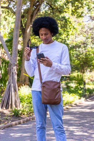 African american man using smartphone and sunglasses on sunlit park path with crossbody bag