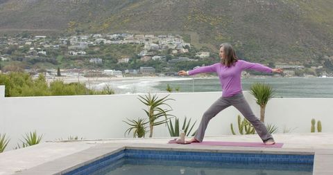 Mature woman practicing yoga on poolside terrace with coastal view and mountain backdrop