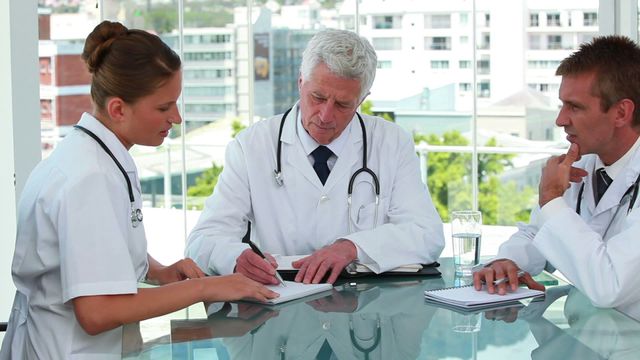 A group of doctors, consisting of an older man and a younger man and woman, are seated around a glass table engaged in intensive discussion. The scene suggests collaboration and decision-making at a hospital or clinic setting. This can be used in contexts related to healthcare teamwork, training, professional development, and medical decision-making.