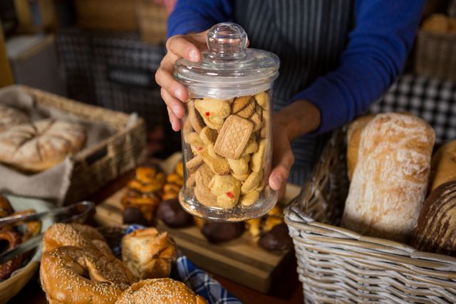 Mid section of staff holding glass jar of cookies at counter in bakery shop