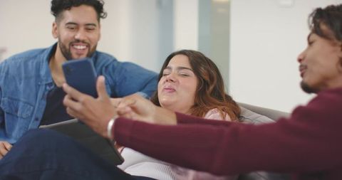Diverse friends laughing and sharing smartphone on modern couch, casual home moment