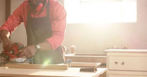 Carpenter Sanding Wood Plank with Power Sander in Sunlit Workshop