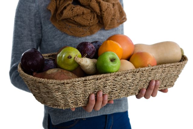 Mid section of woman carrying vegetables and fruits in wicker basket while standing against white background