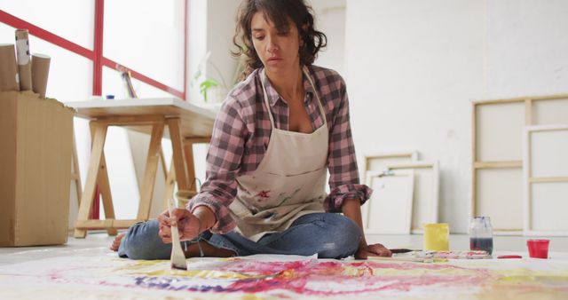 Female artist working on abstract art on canvas in studio. She is sitting down, focused on her work, using various colors. Perfect for use in articles about artistic processes, studio environments, or creativity. Great for promoting art workshops and painting classes.