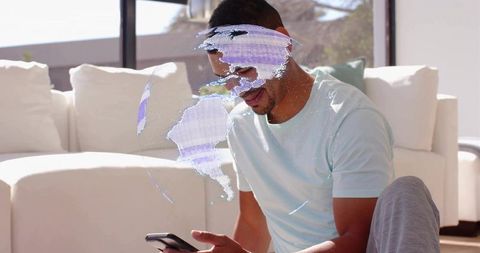 Young man using smartphone with holographic globe projection in sunlit modern living room