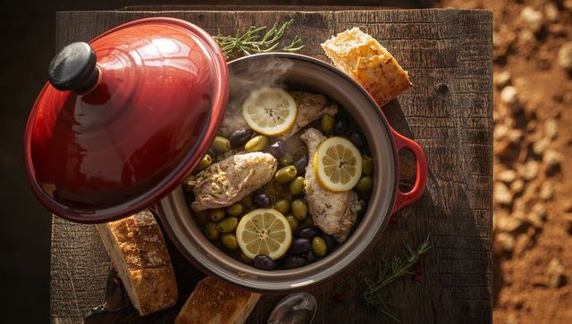 Steaming lemon chicken with green and black olives in red cast-iron pot on rustic table