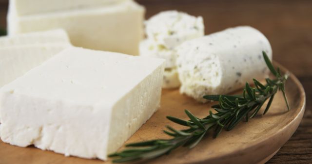 A close-up of various fresh cheeses placed on a rustic wooden board, accompanied by a sprig of rosemary. Perfect for use in culinary articles, food and lifestyle blogs, or restaurant menus. Emphasizes freshness, simplicity, and gourmet dining elements.