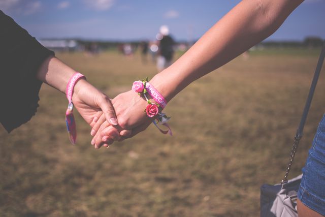 Two women hold hands outdoors with bracelets featuring pink flowers, signifying friendship and bond. Suitable for use in articles about friendship, unity, support groups, and outdoor activities promoting well-being and connection.