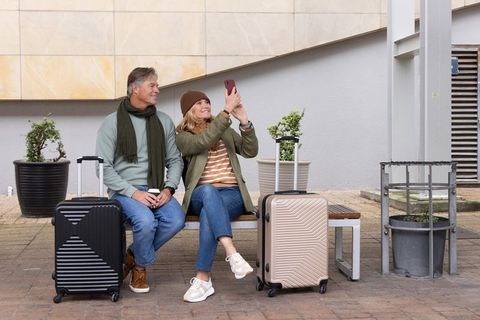 Middle-aged couple taking selfie while waiting with rolling suitcases on urban bench